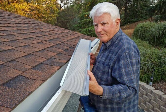 An older man in a plaid shirt stands on a ladder next to a house roof, placing a Gutterglove gutter guard into the rain gutter, demonstrating its easy installation, with trees in the background.