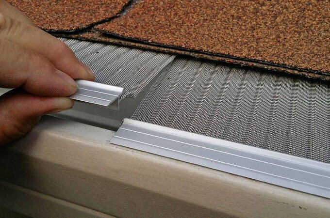 A hand installs a stainless steel micro-mesh gutter guard under roof shingles, securing it along the gutter edge to help prevent debris from entering.