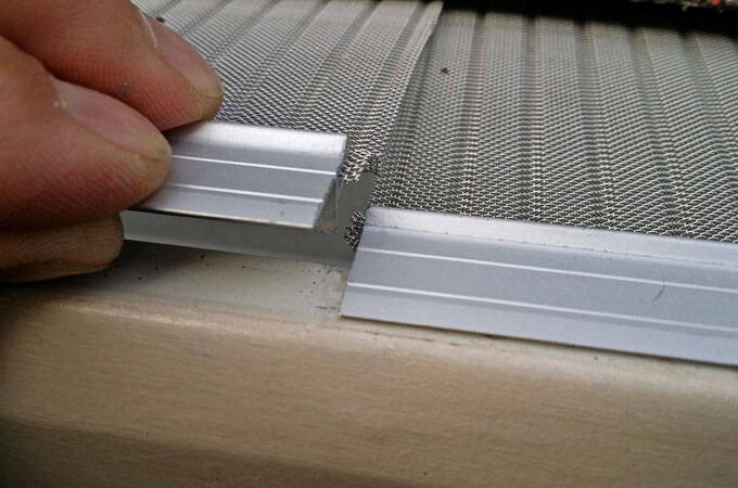 A close-up of a person’s hand aligning two pieces of silver frame over a stainless steel micro-mesh screen, likely assembling or repairing a window or door screen with Gutterglove.
