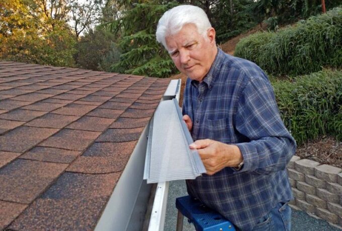 An older man in a plaid shirt stands on a ladder, installing a Gutterglove gutter guard on the edge of a roof, surrounded by trees and greenery, demonstrating its easy installation.