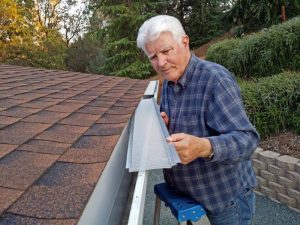 An older man in a plaid shirt stands on a ladder next to a house, installing a mesh gutter guard along the roof edge. Trees and a garden wall are visible in the background.