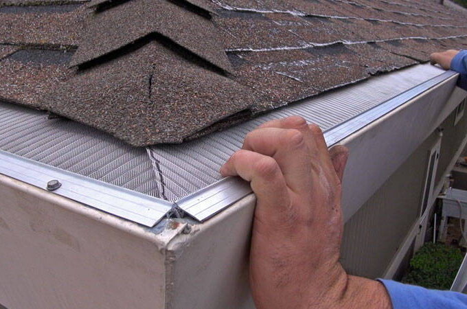A person installs a stainless steel micro-mesh gutter guard, like Gutterglove or Easy On Gutter Guard, along the edge of a shingled roof, securing it over the rain gutter with both hands.