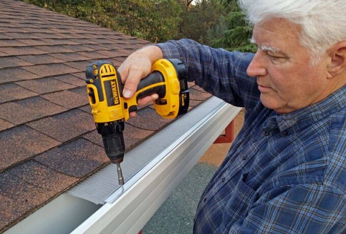 An older man in a plaid shirt uses a yellow cordless drill to install a Gutterglove gutter guard along the edge of a shingled roof. Trees are visible in the background.