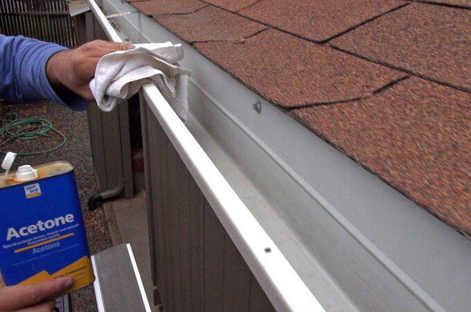 A person cleaning the edge of a house gutter with a cloth and holding a can of acetone, preparing to install Easy On Gutter Guard featuring stainless steel micro-mesh. The roof has brown shingles, and a yard with a green hose is visible in the background.