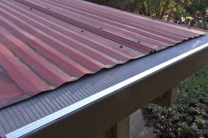 A metal roof with red panels and an Easy On Gutter Guard installed along the edge, surrounded by greenery and trees in the background.