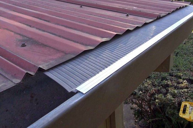 A close-up view of a metal roof with an Easy On Gutter Guard featuring stainless steel micro-mesh installed along the edge to prevent debris from entering the gutter. Bushes and a yellow ladder are visible on the ground below.