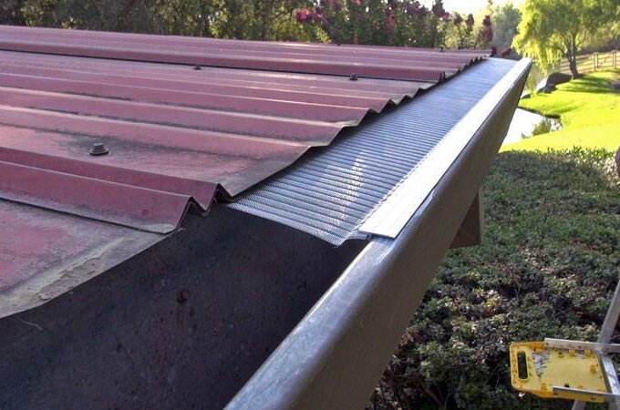 A close-up of a red metal roof with a Gutterglove stainless steel micro-mesh gutter guard installed, viewed from the edge. A yellow step ladder stands beside the roof, with greenery and trees visible in the background.