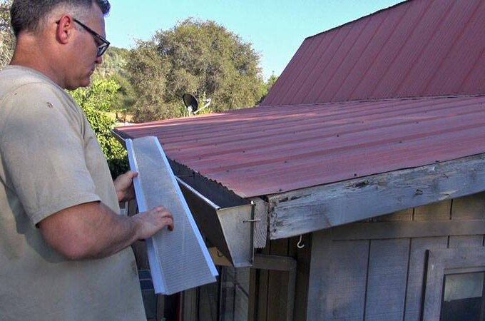 A man holds a section of stainless steel micro-mesh gutter guard, preparing for an easy installation along the edge of a red metal roof on a wooden building, with trees and blue sky in the background.