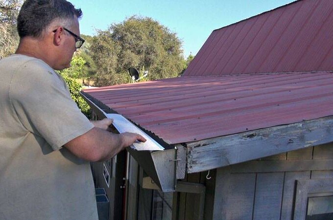 A person wearing glasses measures the edge of a red metal roof on a wooden building, possibly preparing to install Gutterglove gutter guard, with trees and blue sky in the background.