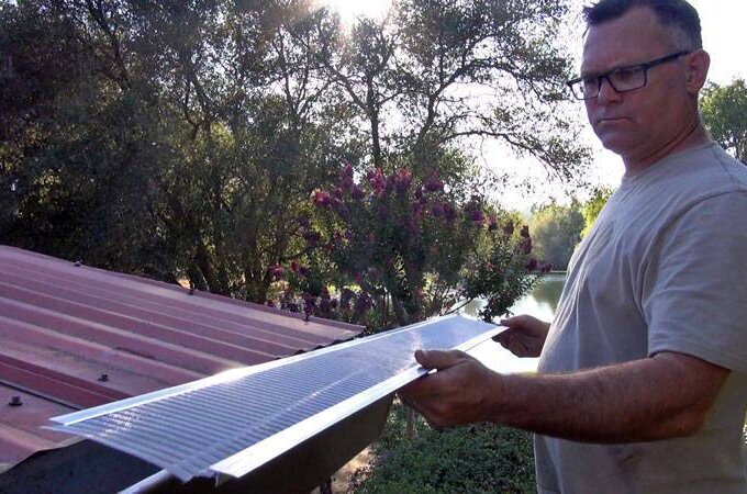 A man wearing glasses and a beige shirt stands by a red metal roof, holding a stainless steel micro-mesh gutter guard outdoors with trees and sunlight in the background, showcasing its easy installation.