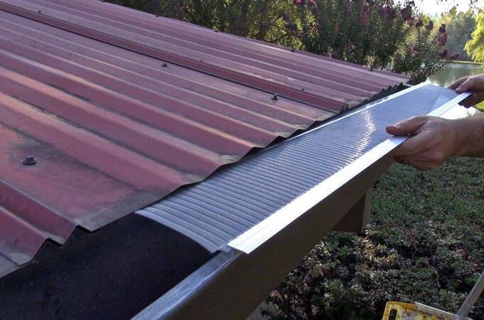 A person installs a Gutterglove metal mesh gutter guard along the edge of a sloped red metal roof above a gutter, surrounded by greenery.