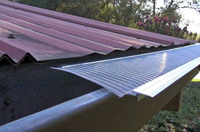 A person installs a stainless steel micro-mesh gutter guard along the edge of a corrugated red metal roof to protect the rain gutter from debris. Trees and greenery are visible in the background.