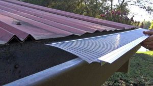 A person installs a metal mesh gutter guard under the edge of a red metal roof to cover an open rain gutter, with trees visible in the background.
