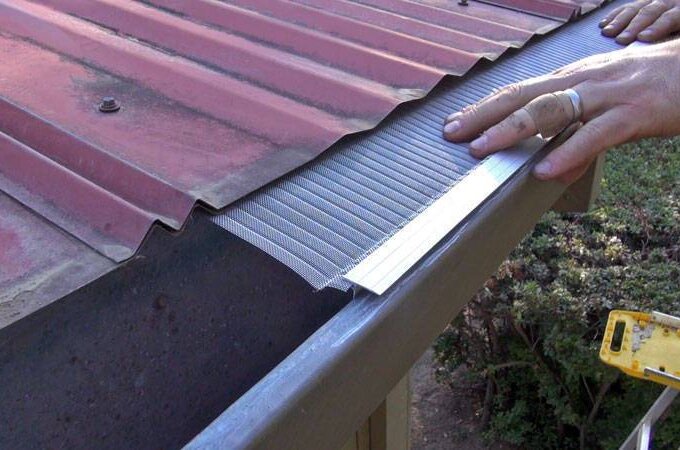 A person installs a Gutterglove gutter guard under the edge of a red metal roof to prevent debris from entering the rain gutter.