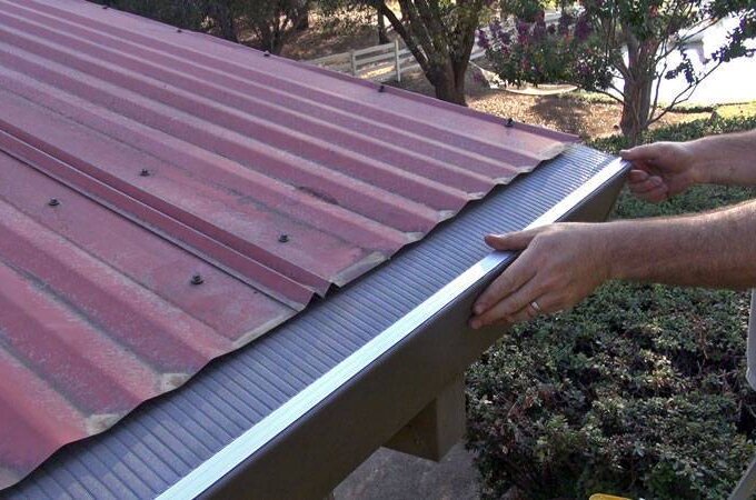 A person installs an Easy On Gutter Guard with easy installation along the edge of a red metal roof to prevent debris from clogging the gutter. Trees and shrubs are visible in the background.
