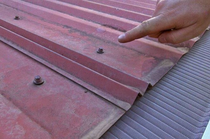 A hand points at a rusty bolt on a red, corrugated metal roof next to a gutter covered with a Gutterglove mesh guard, known for its easy installation.