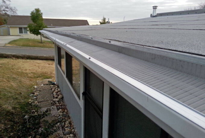 A close-up view of a house roof with a stainless steel micro-mesh gutter guard installed along the edge, preventing leaves and debris from entering. The easy installation is showcased, with the front yard and a nearby street in the background.