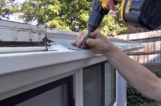A person uses a power drill to install a stainless steel micro-mesh guard over a white rain gutter on a house, ensuring easy installation, with trees and a wooden fence visible in the background.