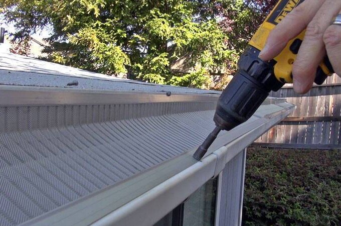 A person uses a power drill to secure a Gutterglove gutter guard onto the edge of a house roof, showcasing its easy installation, with trees and a wooden fence in the background.