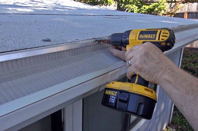 A person uses a yellow and black cordless drill to install a Gutterglove stainless steel micro-mesh gutter guard on the edge of a roof, holding the guard in place while drilling.