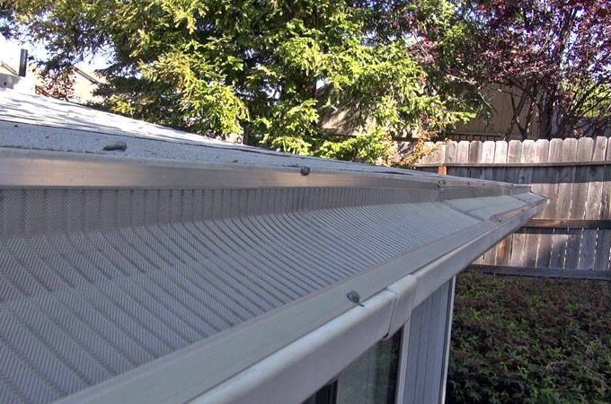 Close-up view of a Gutterglove stainless steel micro-mesh gutter guard installed along the roof edge, with trees, a wooden fence, and greenery in the background.