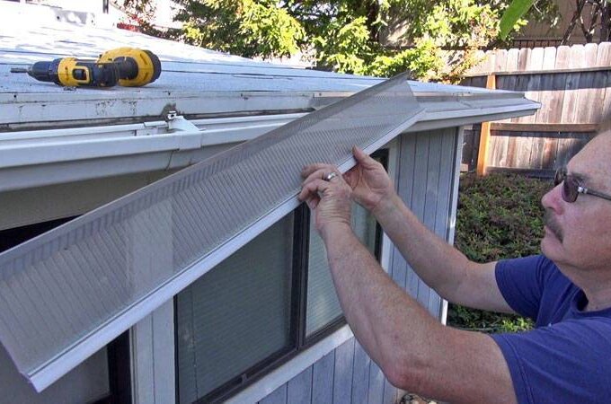 A man installs a Gutterglove gutter guard on a house roof, holding the guard above the gutter for easy installation. A yellow power drill rests on the roof, with trees and a wooden fence visible in the background.