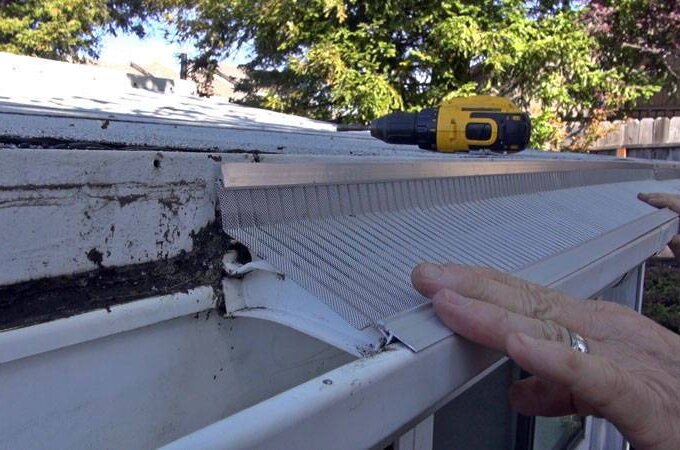A person installs a stainless steel micro-mesh gutter guard on a roof edge, pressing it into place with one hand. A yellow cordless drill rests nearby, and trees are visible in the background—perfect for Costco shoppers.