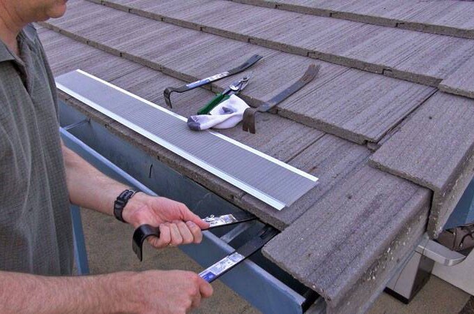 A person installing an Easy On Gutter Guard under the edge of a tiled roof, using hand tools like pliers and a pry bar, with additional tools and materials placed nearby on the roof.