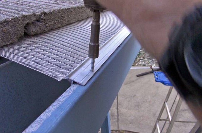 A person uses a power tool to install a mesh gutter guard on the edge of a roof, securing it with a screw. Part of a concrete shingle and blue gutter are visible.