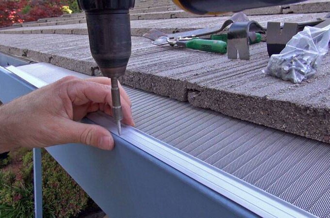 A person uses a power drill to install a metal mesh gutter guard on a roof edge, with tools and screws visible on the shingles nearby.