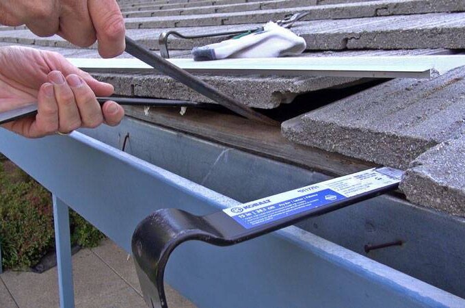A person uses a tool to install a metal drip edge along the edge of a sloped roof with gray tiles, above a blue gutter featuring an Easy On Gutter Guard. Protective gloves and other tools rest on the nearby tiles.