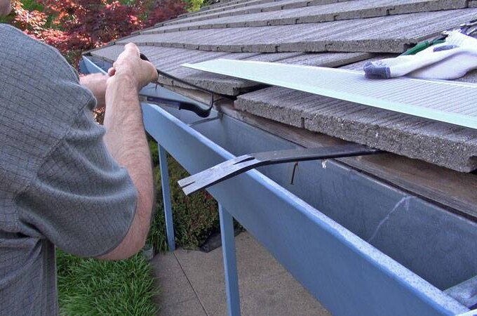 A person is cleaning or repairing a roof gutter with tools next to a house with a tile roof. Gutterglove and work gloves are placed nearby, highlighting easy installation. Green grass and shrubs are visible below.