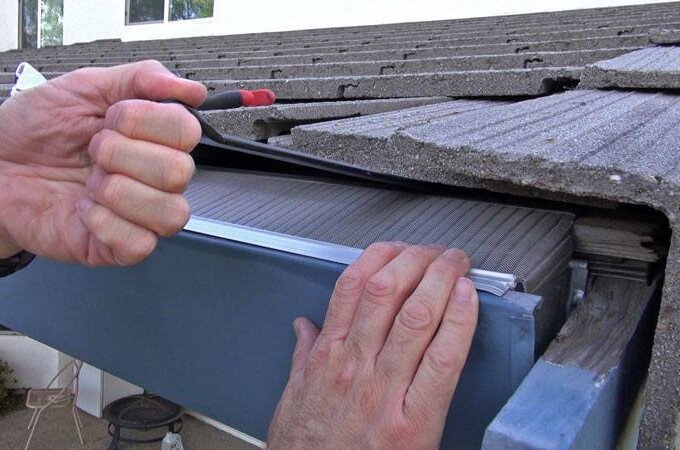 Close-up of a person using a screwdriver to install or repair a mesh screen under roof tiles and above a blue rain gutter on a house.