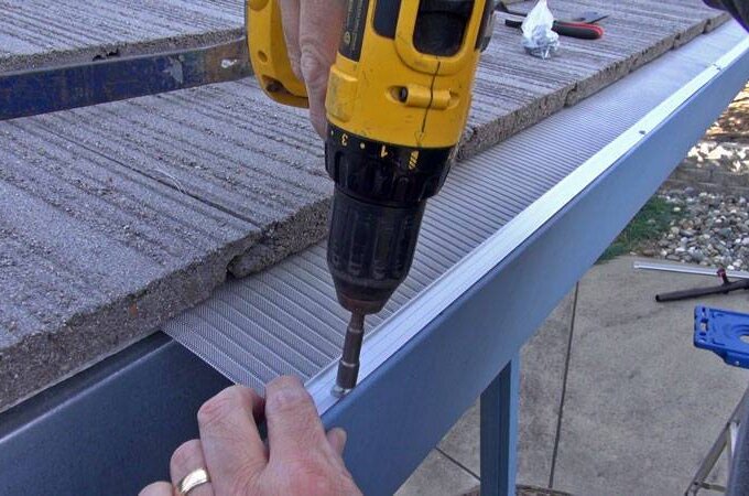 A person uses a yellow power drill to secure a metal gutter guard to the edge of a roof, with concrete roof tiles visible above the gutter.