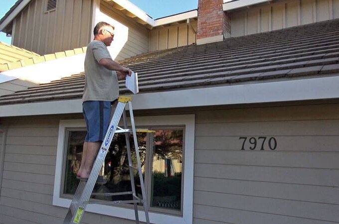 A man standing on a ladder is cleaning or inspecting the gutter on the roof of a beige house with the number 7970 beside a window, demonstrating how Gutterglove from Costco offers easy installation for homeowners.