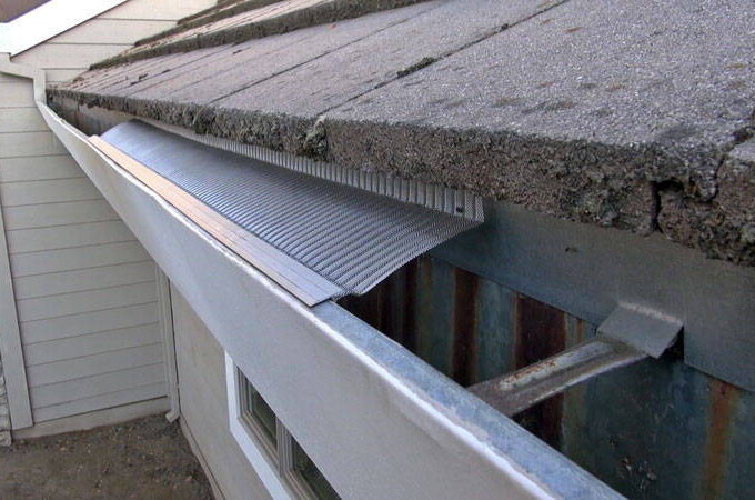 A close-up view of a house roof with a Gutterglove gutter guard partially installed, showing the easy installation above the gutter to prevent debris from entering. The roof has asphalt shingles and light-colored siding.