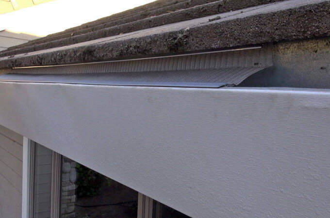 A close-up view of a house roof edge with a metal gutter partially covered by a curved, perforated Easy On Gutter Guard from Costco beneath the roof shingles for easy installation.