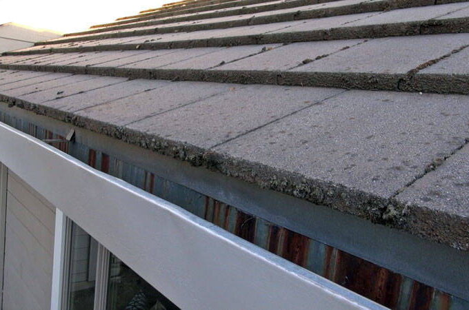 Close-up view of a sloped house roof with gray shingles and a clean, white metal gutter featuring an Easy On Gutter Guard with stainless steel micro-mesh; a portion of the exterior wall and windows is visible below.