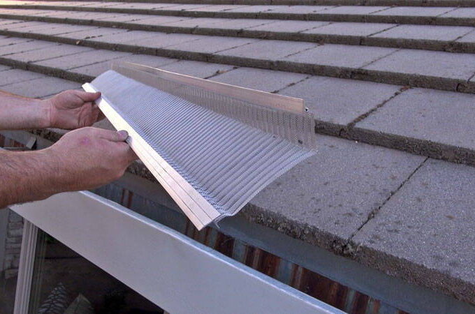 A person holds a Gutterglove stainless steel micro-mesh gutter guard above a house gutter, preparing to install it under the edge of a row of gray roof tiles, similar to those available at Costco.