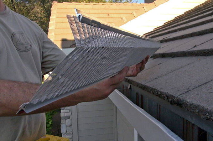 A person installs a stainless steel micro-mesh gutter guard with easy installation along the roof edge to prevent debris from entering the gutter. The house and roof shingles are visible in the background.