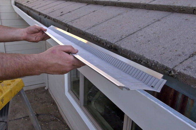 A person stands on a ladder installing a Gutterglove gutter guard along the edge of a house roof to prevent debris from clogging the rain gutter with easy installation.