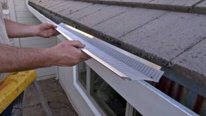 A person installs a metal mesh gutter guard on a house roof to prevent debris from entering the gutter.