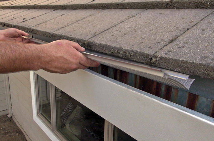 A person installs a piece of metal flashing under the shingles at the roof edge above a rain gutter, showing easy installation to protect against water damage. The hands position the flashing for added protection with Easy On Gutter Guard.