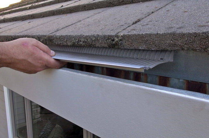 A person installs a Gutterglove gutter guard under the edge of a shingled roof to prevent debris from clogging the rain gutter below, similar to those available at Costco.