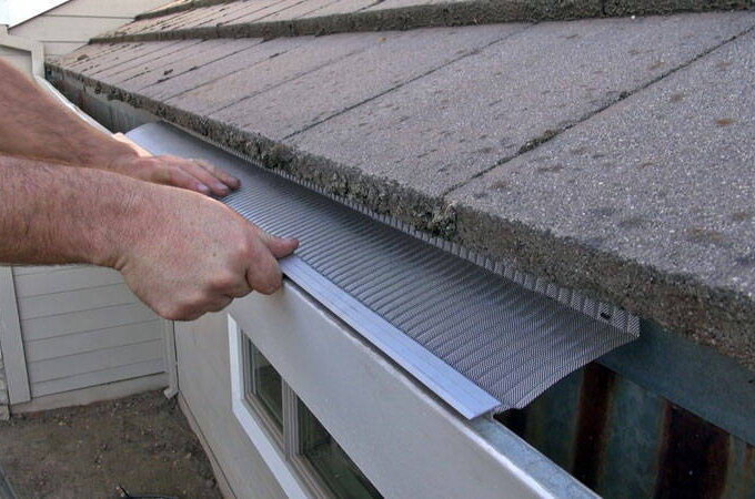A person installs a Gutterglove stainless steel micro-mesh gutter guard along the edge of a roof, using their hands to hold the guard in place under the shingles to prevent debris from entering the gutter.