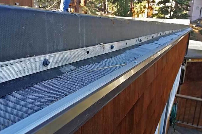Close-up of a house roof gutter with an Easy On Gutter Guard mesh installed to prevent leaves and debris from entering. Trees and part of a wooden deck are visible in the background.