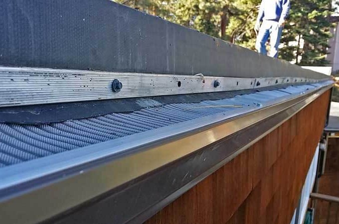 Close-up view of a metal gutter with a Gutterglove gutter guard featuring easy installation on the edge of a roof, with trees and a person in the background.