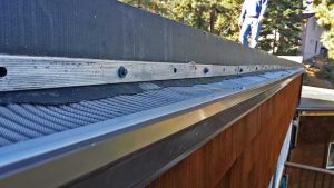 Close-up of a house gutter with a metal mesh guard installed, preventing debris from entering. A person stands on the roof in the background near trees and other houses.