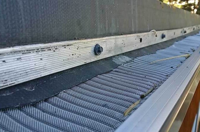 Close-up view of a roof gutter protected by an Easy On Gutter Guard with stainless steel micro-mesh, designed to keep out debris. Screws, dirt, and pine needles are visible on the mesh and along the roof’s edge.