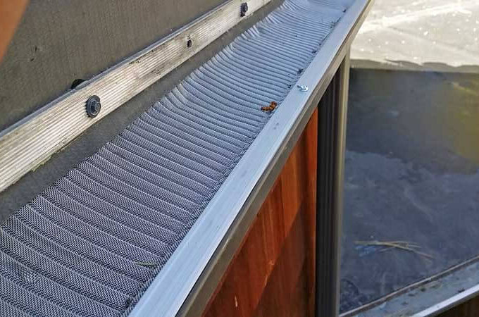 Close-up of a stainless steel micro-mesh Gutterglove gutter guard installed on a roof edge, designed to keep debris out. Some small debris is visible on the mesh screen. Easy installation makes this guard ideal for DIY homeowners.